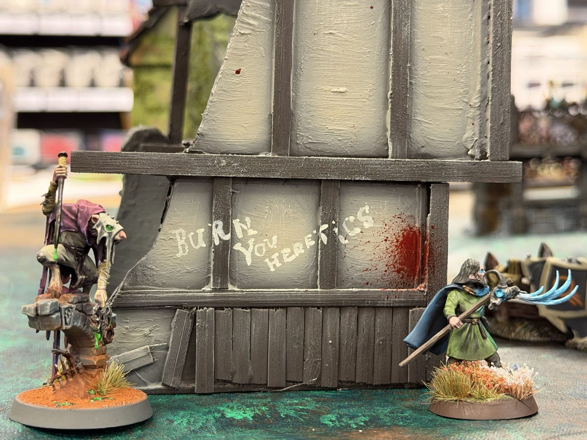 Twee geschilderde miniaturen bij een vakwerkmuur met de tekst 'Burn you heretics' en een bloedvlek