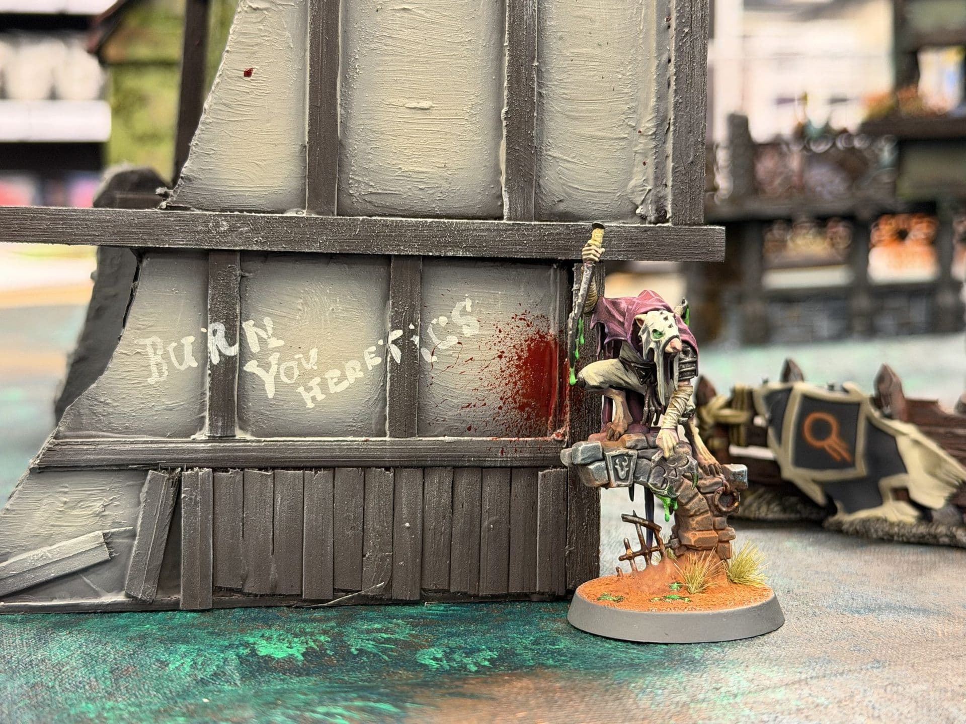 Rattenachtige miniatuur in paarse mantel op een stenen muurtje naast een vakwerkmuur met de tekst 'Burn you heretics'