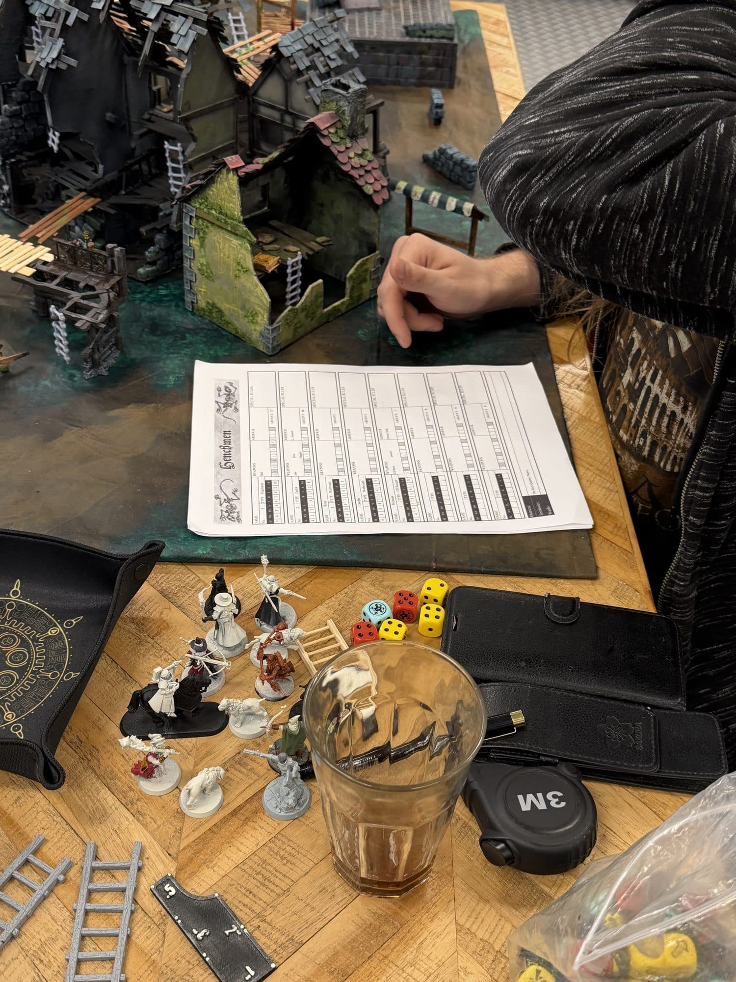 Speler aan tafel met een warbandformulier, dobbelstenen en miniaturen, met de Mordheim tafel op de achtergrond