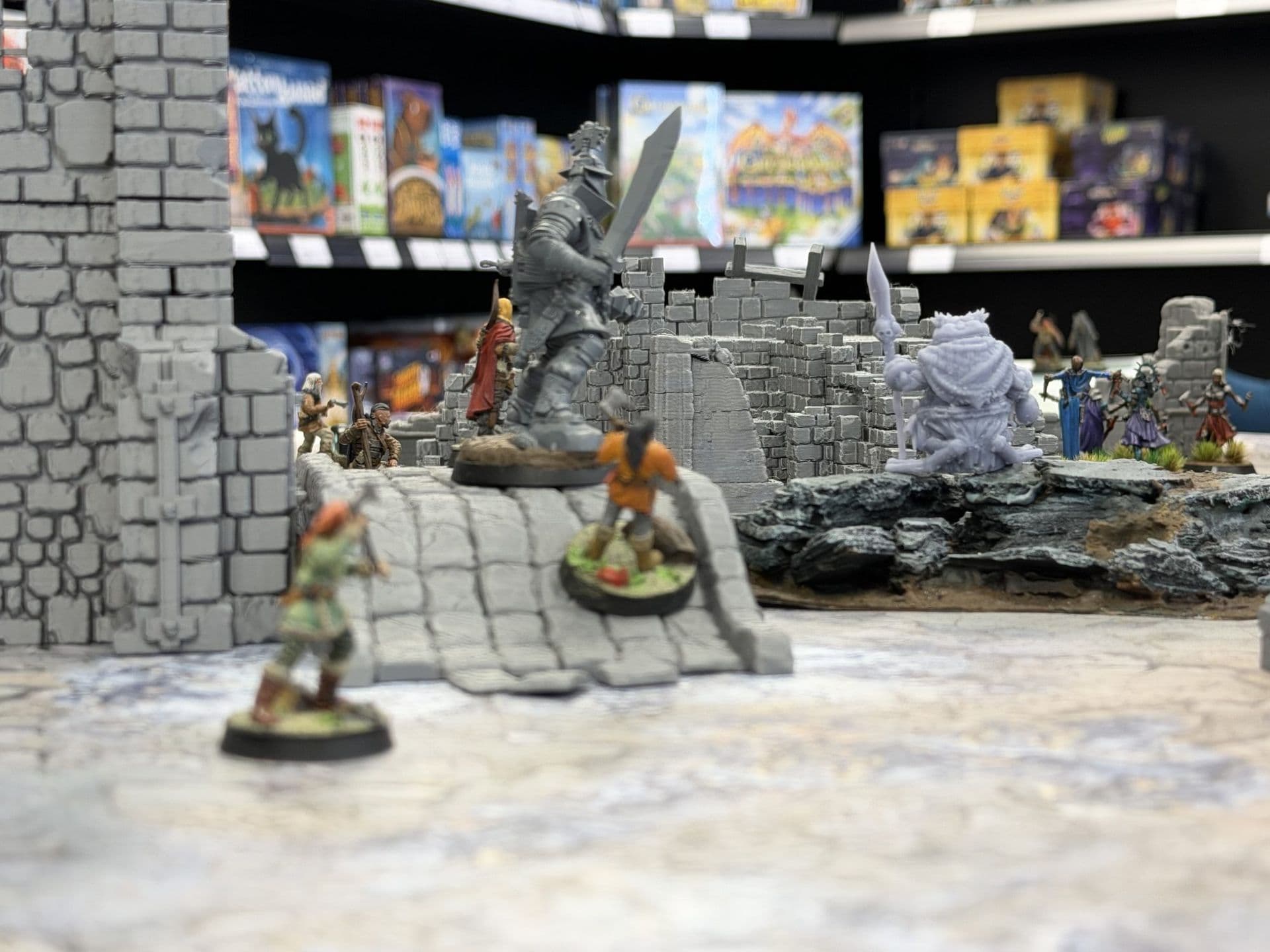 Miniaturen tussen grijze stenen ruïnes en een trap, met een winkelkast vol spellen op de achtergrond