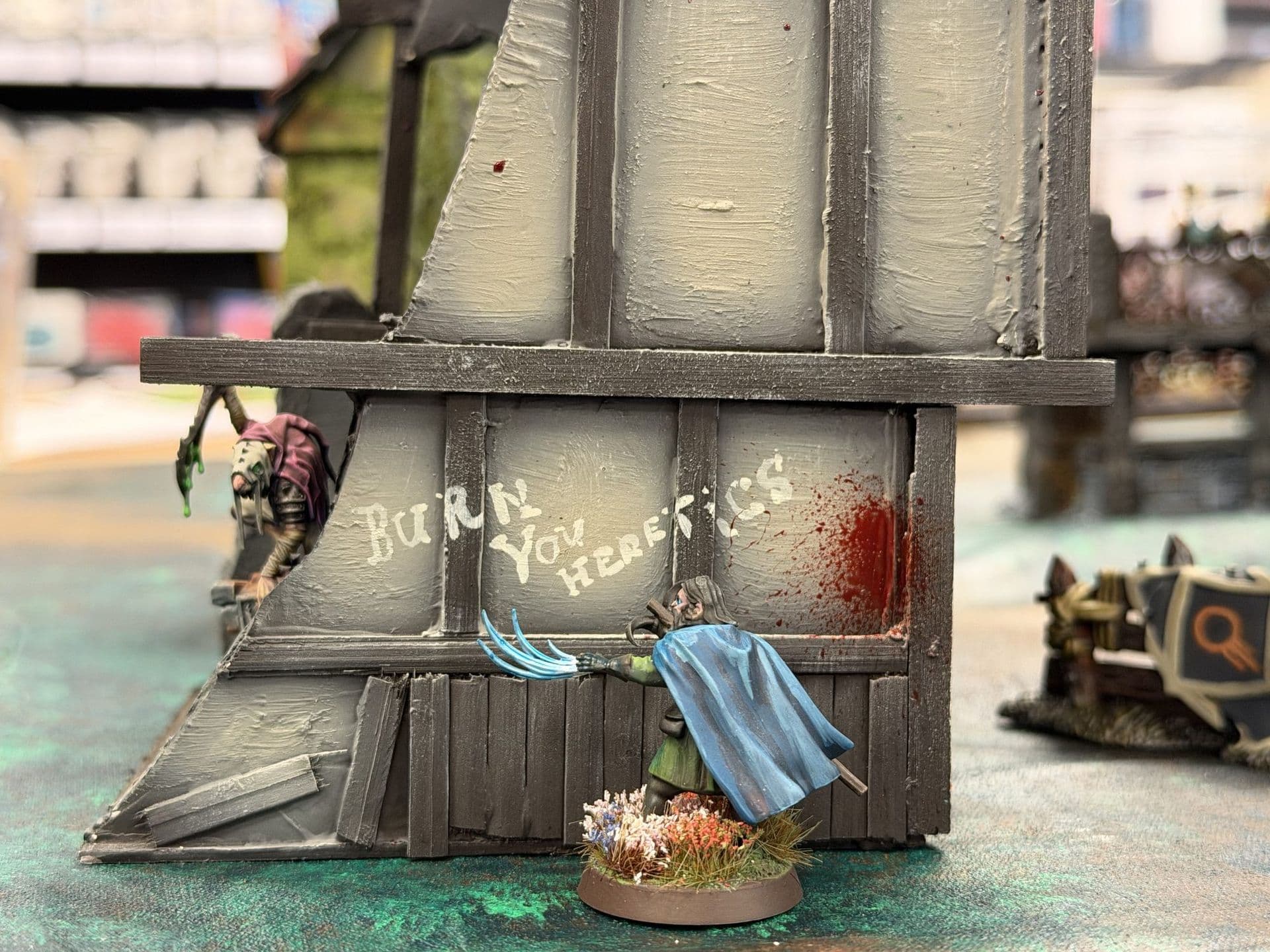Miniatuur in blauwe mantel voor een houten muur met de tekst 'Burn you heretics' en een bloedvlek