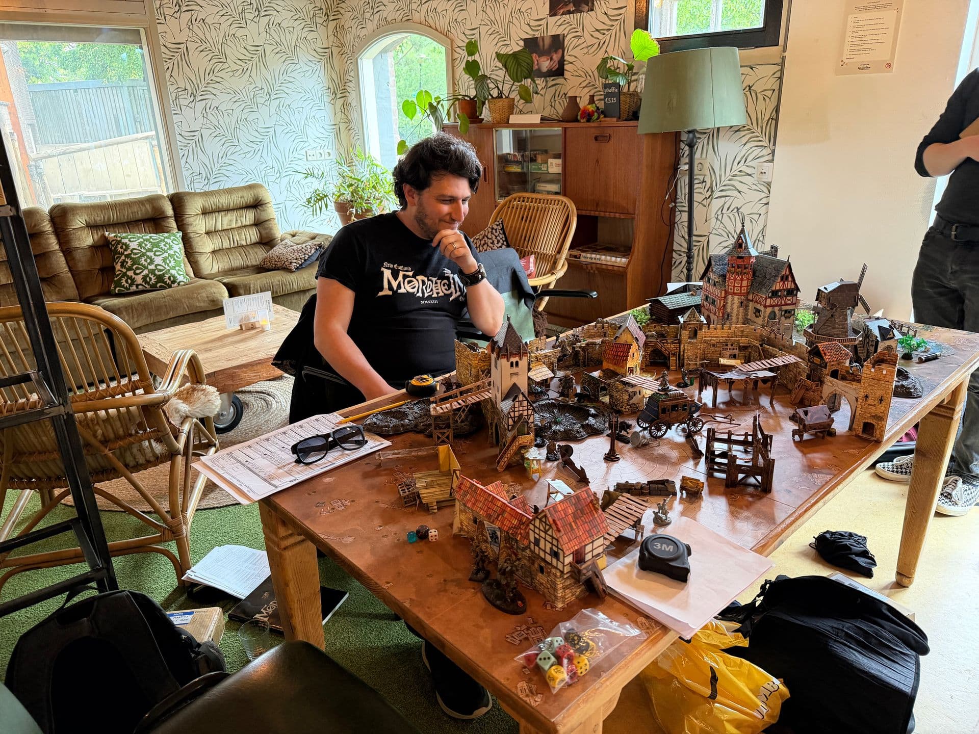 Speler in een Mordheim shirt kijkt naar een speeltafel vol gebouwen en miniaturen