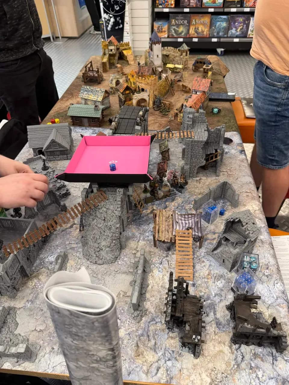 Spelers rond een grote Mordheim speeltafel vol ruïnes en torens, met een roze dobbelbak