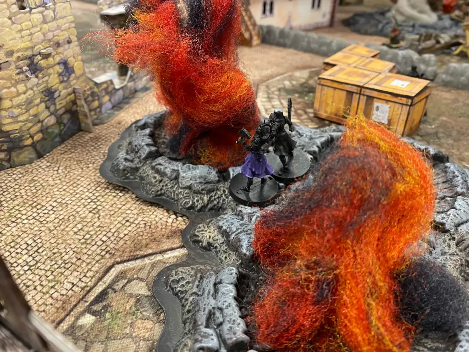 Twee Mordheim miniaturen tussen vuureffecten en rotsen, met stenen huizen en houten kratten op de speeltafel